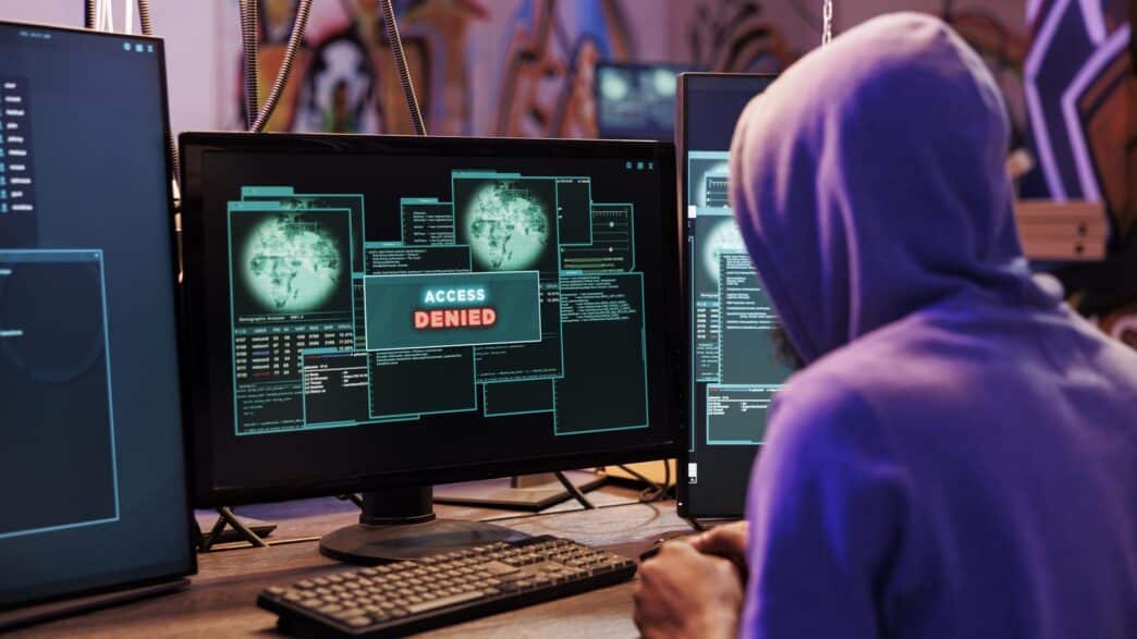 Hacker in a hoodie facing an "Access Denied" alert on computer screens during an attempted cyberattack.