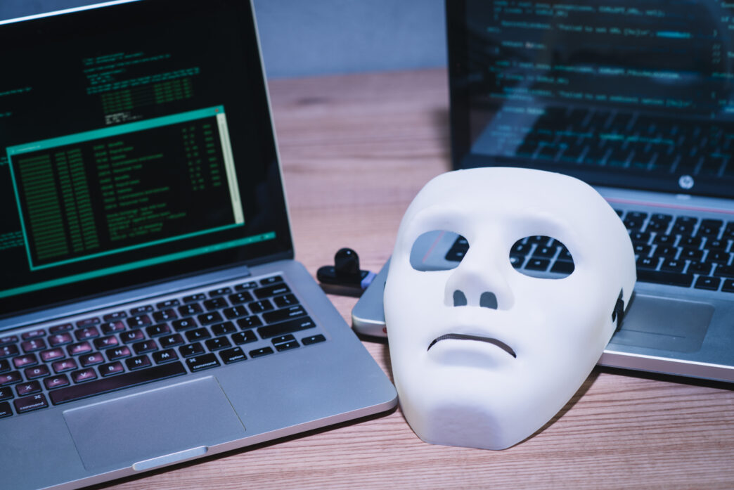 A white face mask resting beside two laptops running code, symbolizing anonymity and cybersecurity.