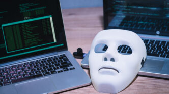 A white face mask resting beside two laptops running code, symbolizing anonymity and cybersecurity.