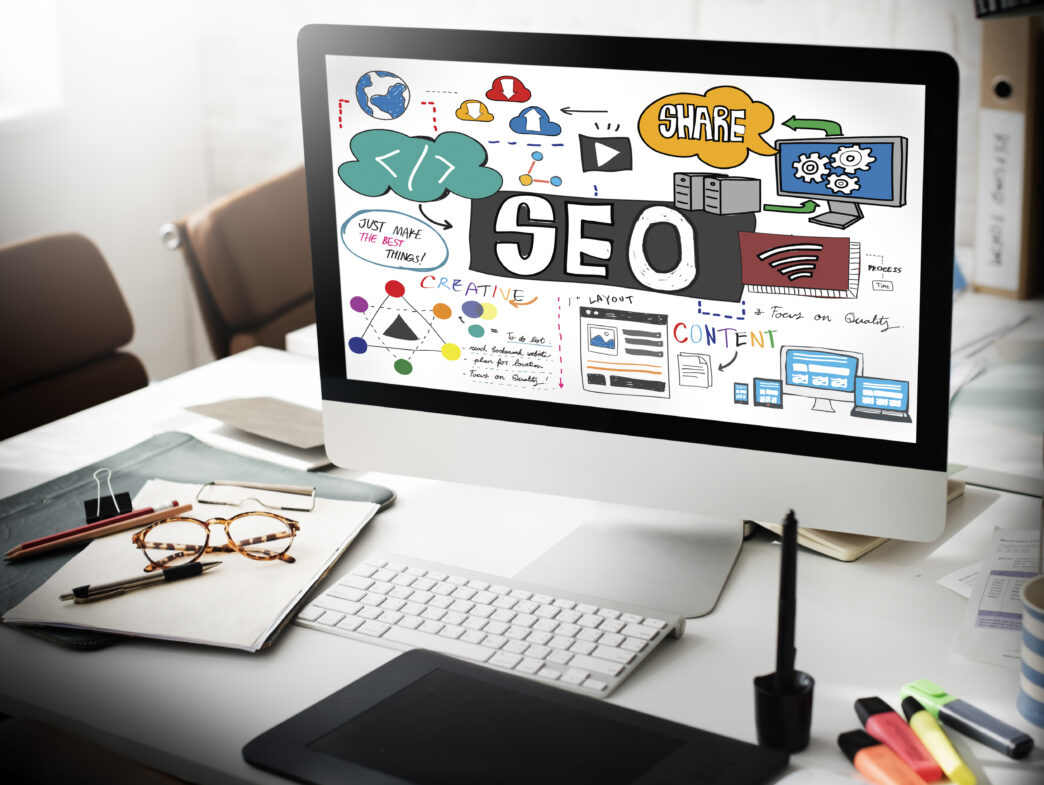 A computer monitor on a desk displaying an SEO concept sketch with colorful icons, text, and diagrams.