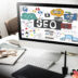 A computer monitor on a desk displaying an SEO concept sketch with colorful icons, text, and diagrams.